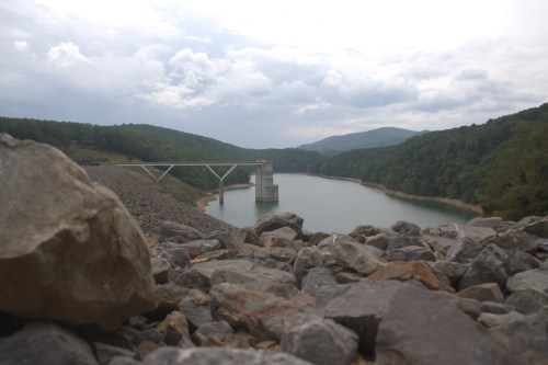 A view of a dam and reservoir surrounded by rocky terrain and lush green hills under a cloudy sky.