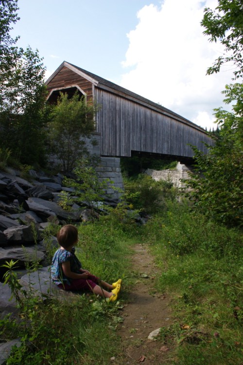 A child sits on a path near a covered wooden bridge surrounded by greenery and rocks under a blue sky.