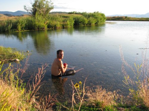 A shirtless man sits in a calm pond surrounded by greenery and mountains in the background, enjoying the water.