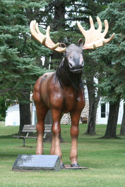 A life-sized moose statue stands on a grassy area, surrounded by trees, with a plaque at its base.