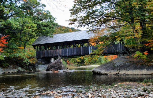 A wooden covered bridge spans a calm river, surrounded by colorful autumn foliage and rocky banks.
