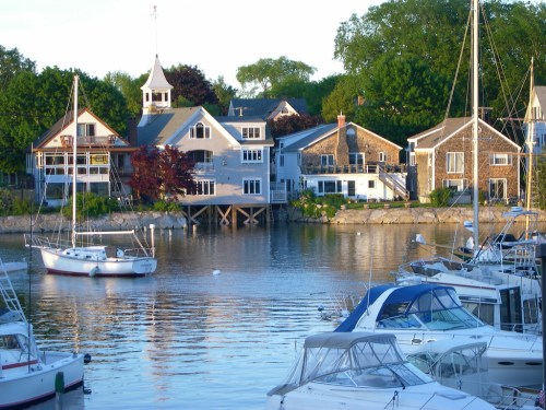 A serene waterfront scene with boats docked and charming houses lining the shore, surrounded by lush greenery.