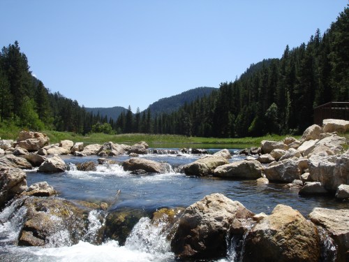 A serene river flows over rocks, surrounded by lush green trees and mountains under a clear blue sky.