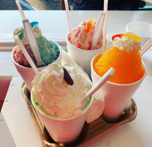 Four colorful snow cones in cups, topped with whipped cream and candy, served in a cardboard tray.