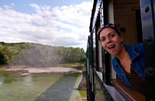 A woman leans out of a train window, smiling, with a scenic river and trees in the background.