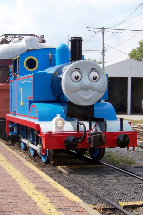 A cheerful blue train with a smiling face, parked on a railway track near a building.