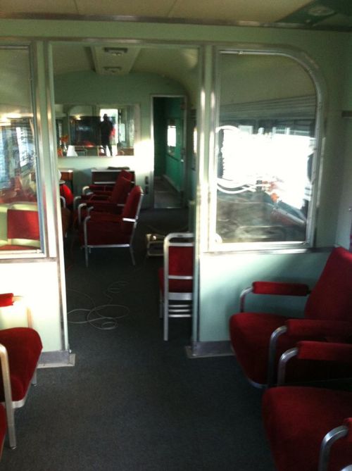 Interior of a vintage train car featuring red upholstered chairs and green walls, with reflections in the windows.