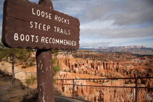 Sign warning of loose rocks and steep trails, with a scenic view of a canyon landscape in the background.