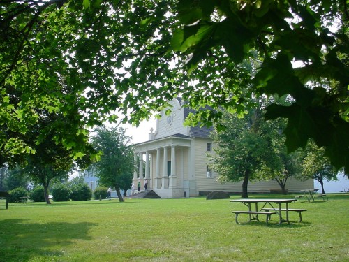 A historic building with columns surrounded by green trees and a grassy area, featuring picnic tables in the foreground.