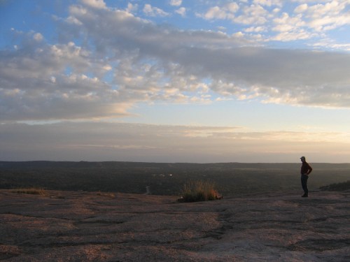 A person stands on a rocky outcrop, gazing at a vast landscape under a cloudy sky during sunset.