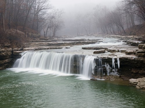 A serene waterfall cascades over rocky ledges into a misty, green river surrounded by bare trees.