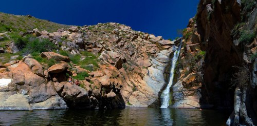 A scenic waterfall cascading down rocky cliffs into a serene pool, surrounded by lush greenery and blue skies.