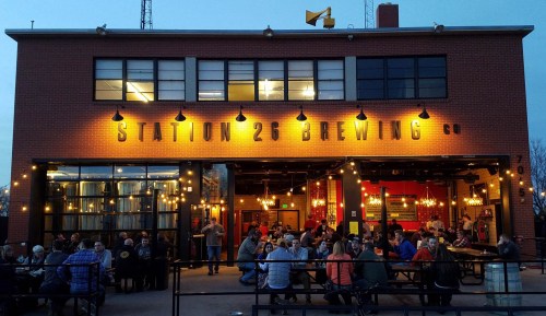 A lively brewery with outdoor seating, string lights, and patrons enjoying drinks at Station 26 Brewing Co. in the evening.