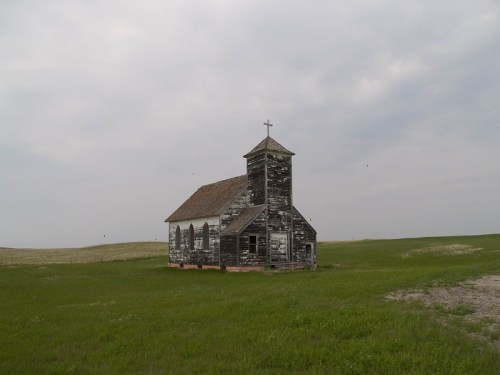 An old, weathered church stands alone in a grassy field under a cloudy sky.