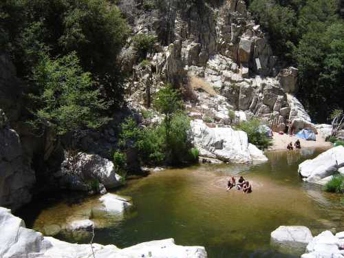 A serene natural pool surrounded by rocky cliffs and greenery, with people relaxing on the shore.