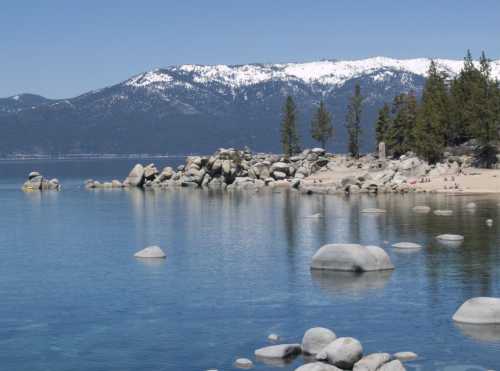 A serene lake scene with clear water, rocky shores, and snow-capped mountains in the background under a blue sky.