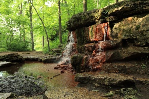 A small waterfall cascades over rocky terrain, surrounded by lush green trees in a serene forest setting.