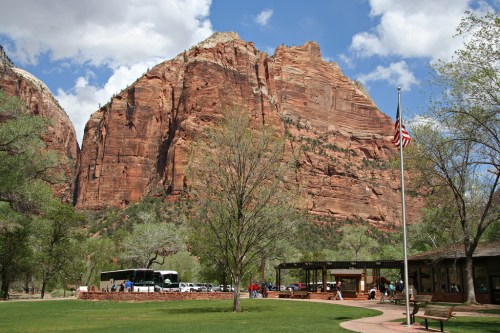 A scenic view of Zion National Park featuring towering red cliffs, a flagpole, and a visitor center with parked vehicles.