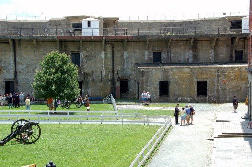 Historic fort with stone walls, grassy courtyard, and visitors exploring. Cannons and trees are visible in the foreground.