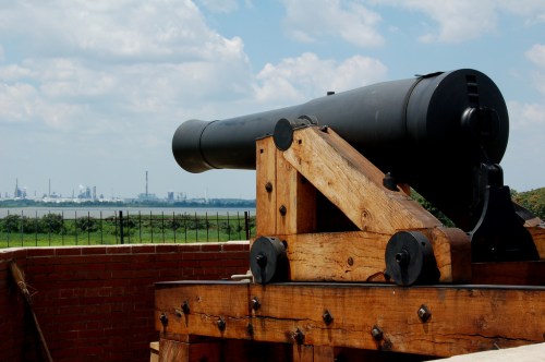 A historic cannon mounted on a wooden base, overlooking a scenic landscape with distant industrial structures.