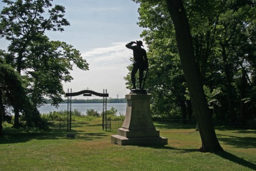 A statue of a soldier stands in a green park, overlooking a lake through a decorative gate surrounded by trees.