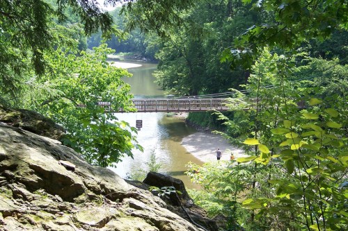 A scenic view of a river with a wooden bridge, surrounded by lush greenery and rocky terrain.