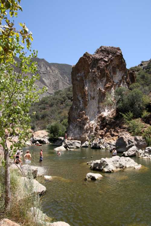 A serene river scene with people swimming near rocky formations and lush greenery under a clear blue sky.