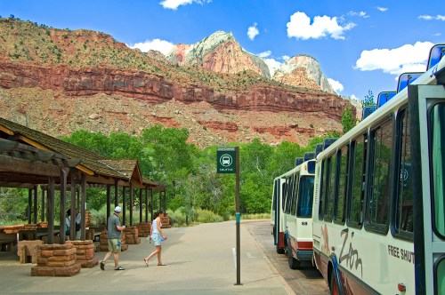 A scenic view of Zion National Park with shuttle buses parked near a stop, surrounded by red rock formations and greenery.