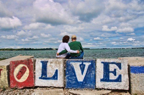 A couple sits on a pier, embracing, with colorful letters spelling "LOVE" in front of them and a cloudy sky above.