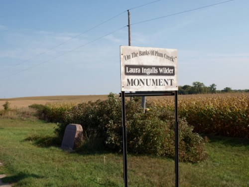 Sign for the Laura Ingalls Wilder Monument, located near a cornfield under a clear blue sky.