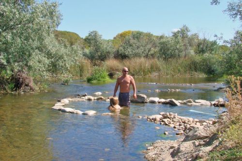 A man stands in a shallow stream while a child plays nearby, surrounded by trees and rocks under a clear blue sky.