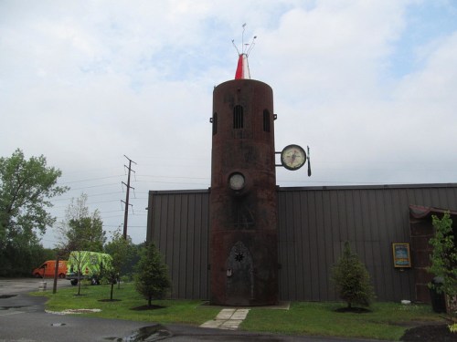 A tall, rusty tower with a red cone on top, featuring gauges and a clock, beside a gray building and green trees.