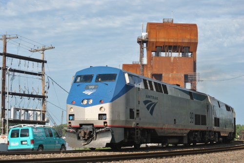 Amtrak train 99 passes by a rusted industrial structure and a teal van on a sunny day.