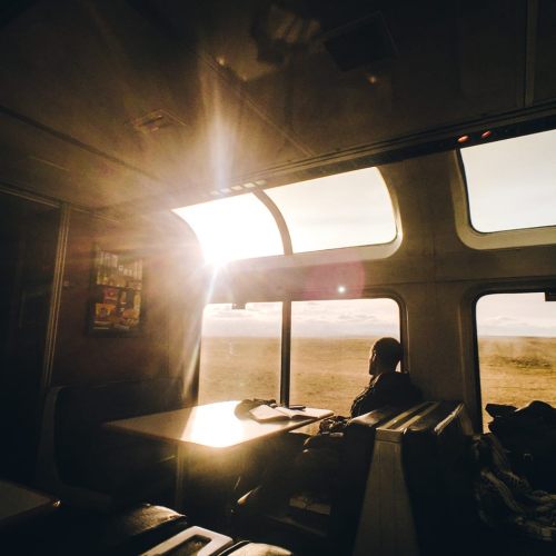 A sunlit train interior with a passenger sitting at a table, gazing out the window at a scenic landscape.