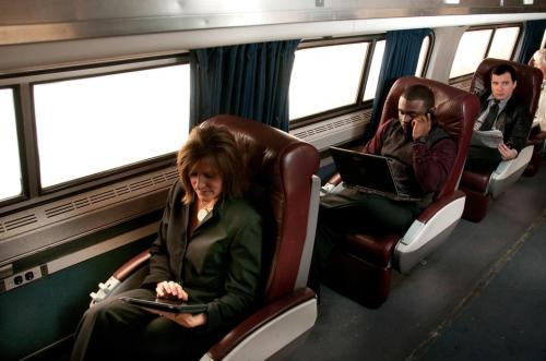 Three passengers in a train car, two using laptops and one on a phone, seated in comfortable leather chairs.