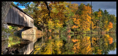 A serene autumn scene featuring a covered bridge over a calm lake, surrounded by vibrant fall foliage reflecting in the water.