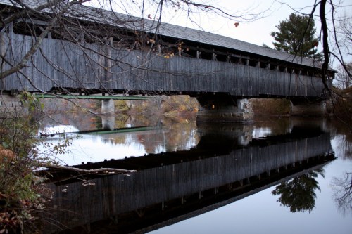 A wooden covered bridge spans a calm river, reflecting its image in the water below, surrounded by autumn foliage.