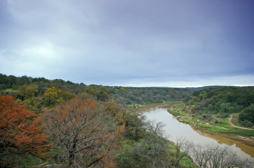 A scenic view of a winding river surrounded by lush greenery and autumn-colored trees under a cloudy sky.
