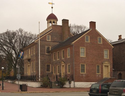 Historic brick building with a dome and weather vane, surrounded by trees and parked cars. Overcast sky.