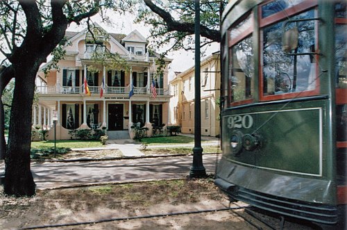 A historic house with flags, surrounded by trees, as a green streetcar passes by in the foreground.