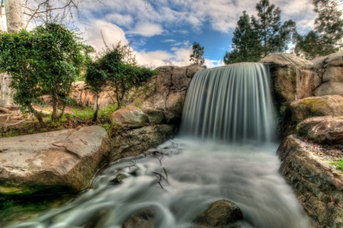 A serene waterfall cascades over rocks, surrounded by lush greenery and a cloudy blue sky.