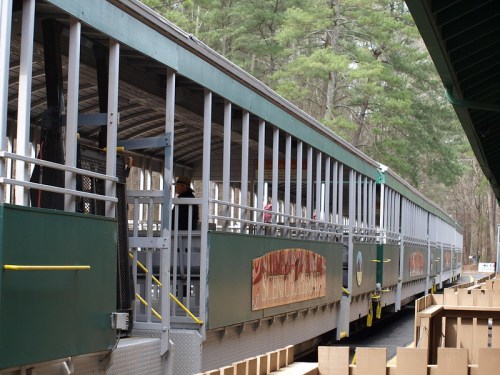 A long, open-air train car parked at a station, surrounded by trees and wooden structures.