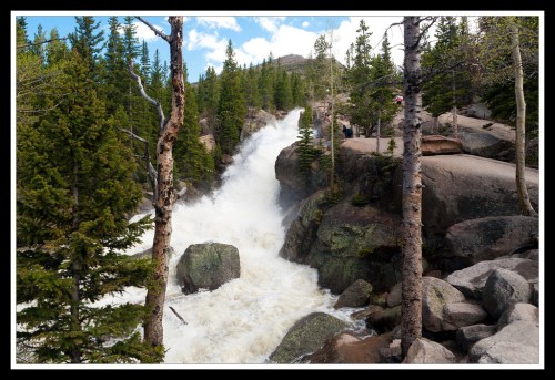 A powerful waterfall cascades over rocks, surrounded by lush green trees and a clear blue sky.