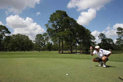 A golfer crouches on the green, focusing on a putt, with trees and a blue sky in the background.
