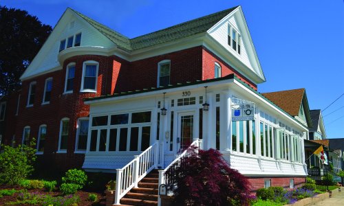A large, charming brick house with a white porch and large windows, surrounded by greenery under a clear blue sky.