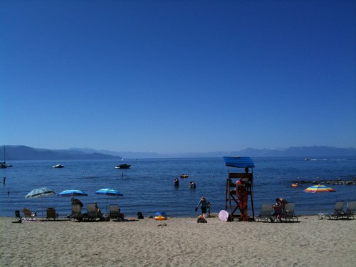 A sunny beach scene with umbrellas, lounge chairs, and people enjoying the water under a clear blue sky.