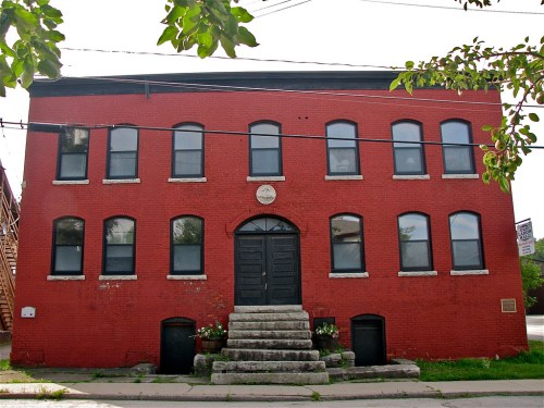 A red brick building with multiple windows and a central black door, featuring stone steps and greenery at the entrance.