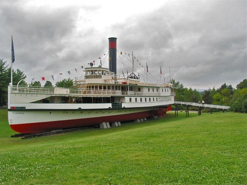 A historic steamboat docked on land, surrounded by green grass and under a cloudy sky, with colorful flags displayed.