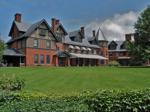 A large, historic brick mansion with multiple gables and a green lawn under a partly cloudy sky.