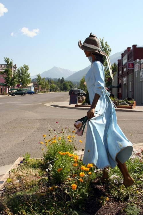 A statue of a woman in a blue dress walking through a flower garden on a sunny street with mountains in the background.